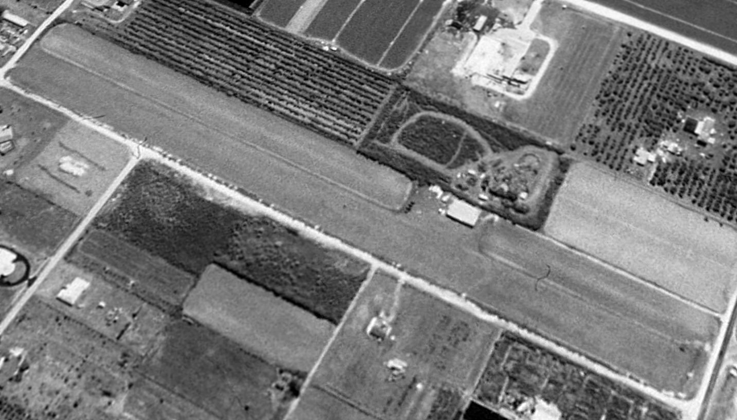 1994 USGS black-and-white aerial of Miami Gliderport looking southeast: a single grass east-west runway with one small hangar on the south side, surrounded by tomato fields and orchards